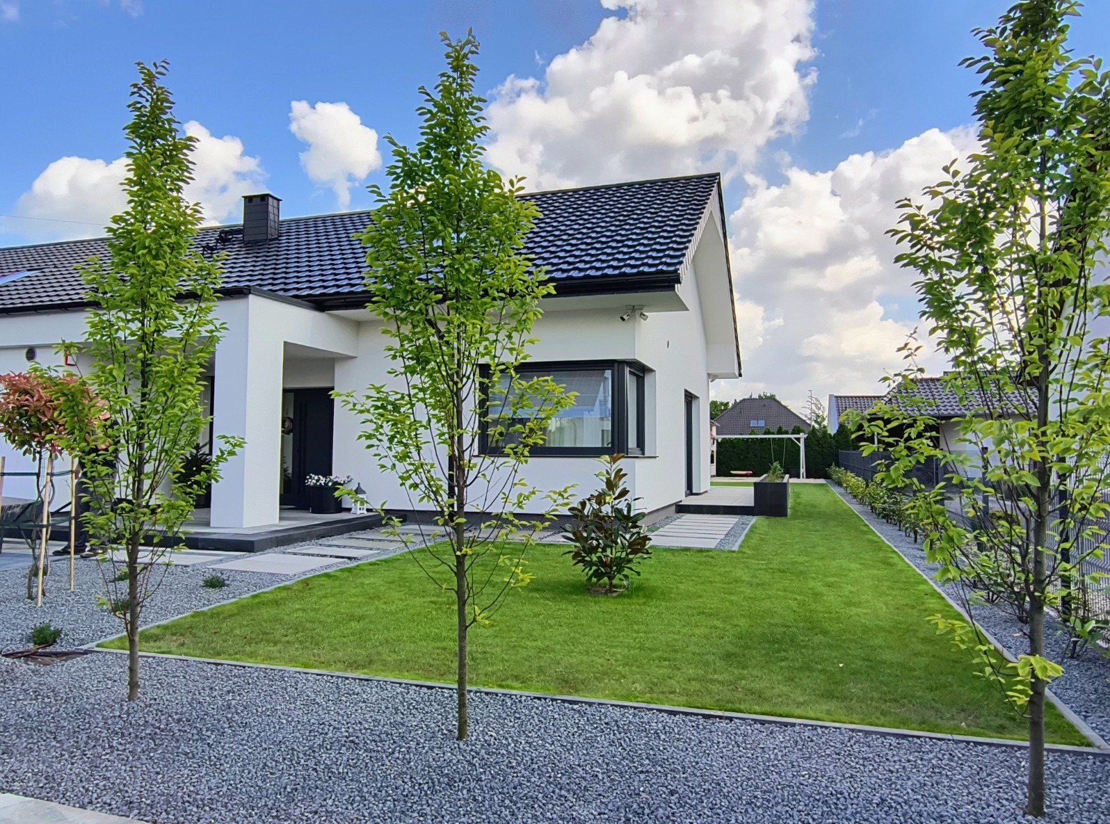Nowoczesny ogród z równo przystrzyżonym trawnikiem, młodymi drzewami i szarą ścieżką z kamienia, otaczający biały dom z czarnym dachem w słoneczny dzień.