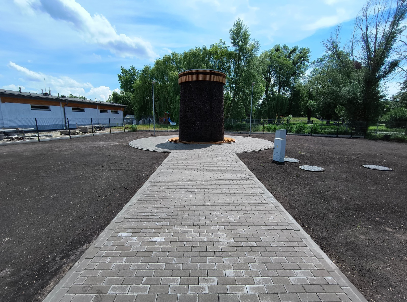Solankowa wieża tężniowa otoczona nową kostką brukową i przygotowanym terenem pod zieleń, w tle budynek i drzewa w słoneczny dzień.
