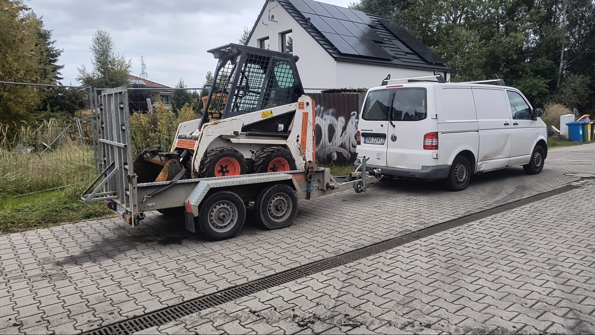 Bobcat na przyczepie, ciągnięty przez białego busa na kostce brukowej. W tle dom z panelami słonecznymi i ogrodzenie.