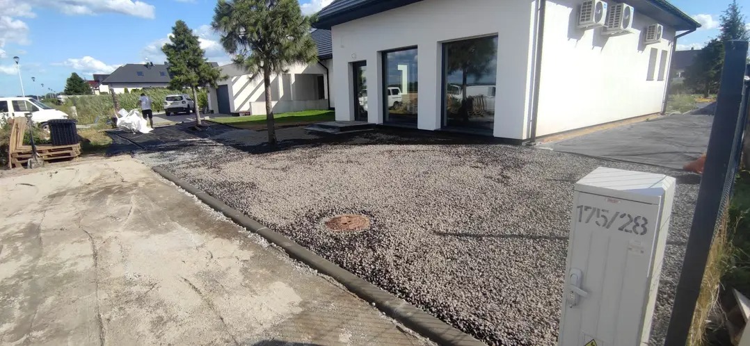 Wysypany żwir na posesji przed nowoczesnym domem z dużymi oknami. Widoczne obrzeża, trawa pod żwirem i pracownik w tle. Częściowo utwardzony teren z lewej.