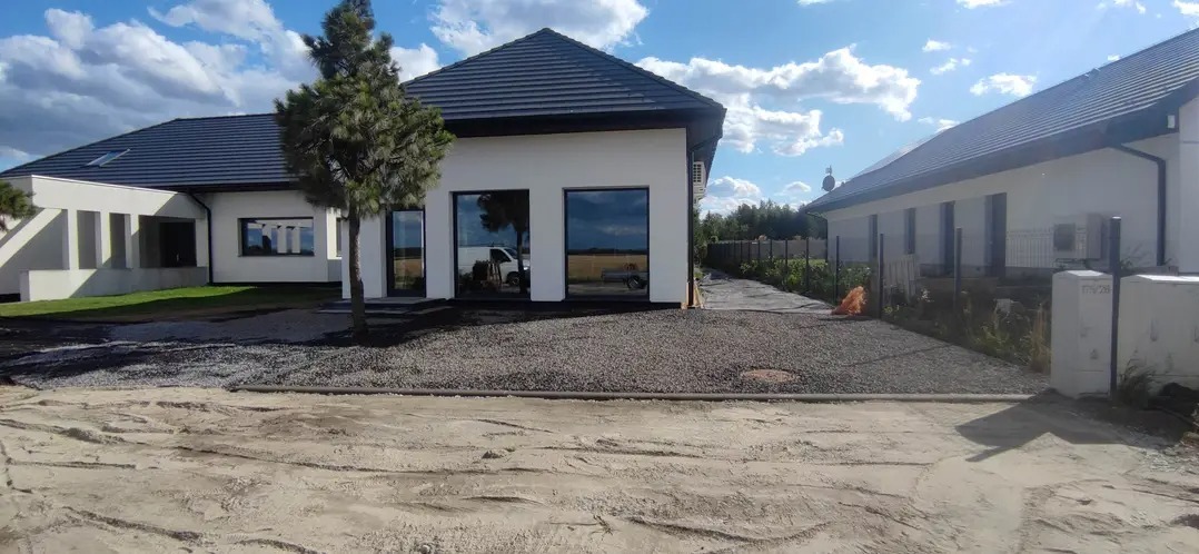 Nowoczesny dom z szarym podłożem z kamienia i młodym drzewkiem iglastym. W oknach odbija się biały samochód dostawczy. Błękitne niebo z chmurami w tle.