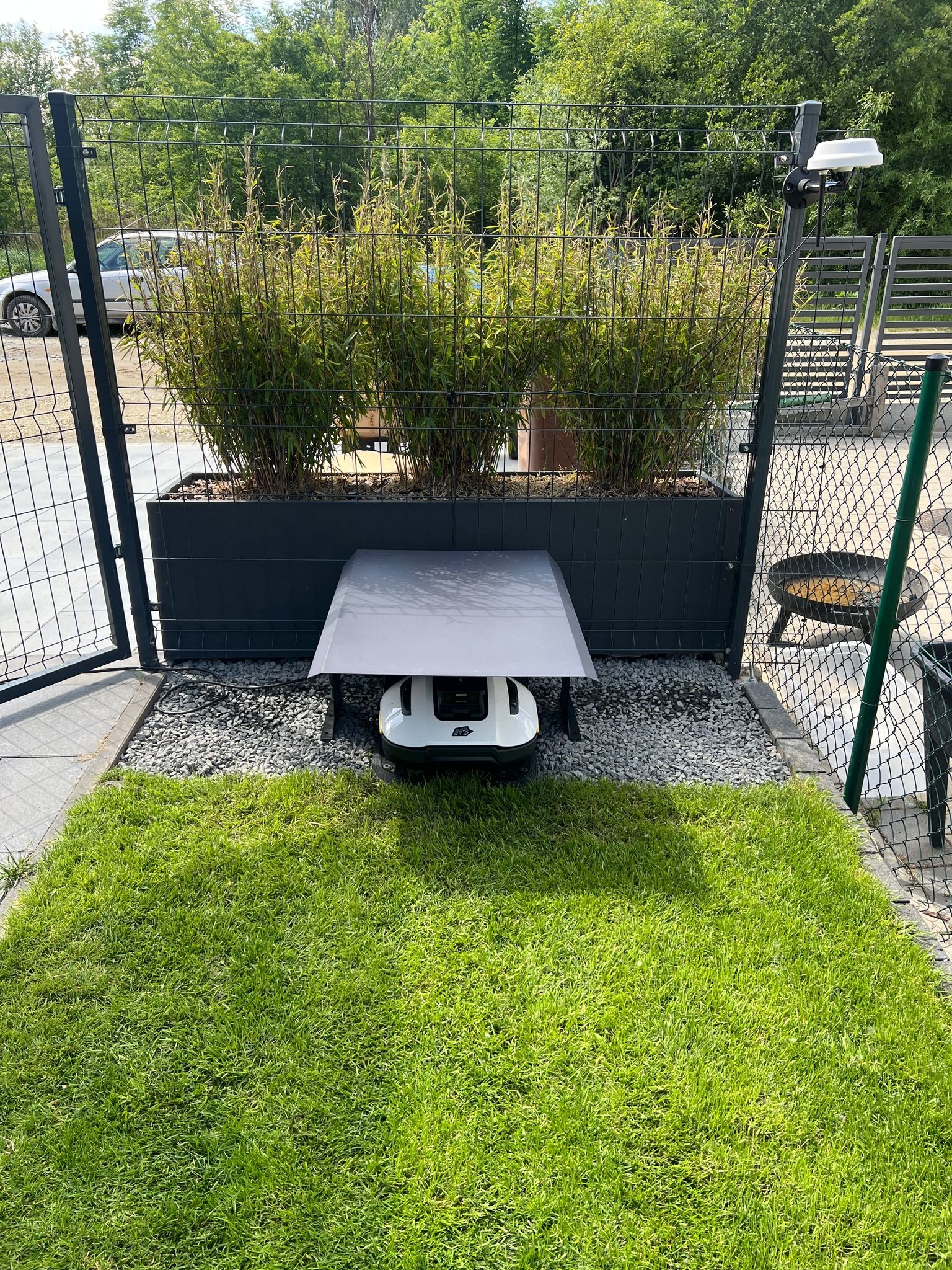 Robot koszący pod zadaszeniem w nowoczesnym ogrodzie z trawą i roślinami w donicy, widok z góry, ogrodzenie panelowe w tle.