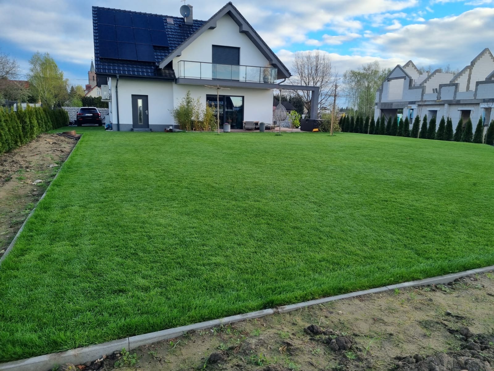Perfekcyjny trawnik z rolki w Namysłowie przed nowoczesnym domem z panelami słonecznymi. Równa, zielona powierzchnia, obrzeża z kostki. Tuje wzdłuż ogrodzenia.