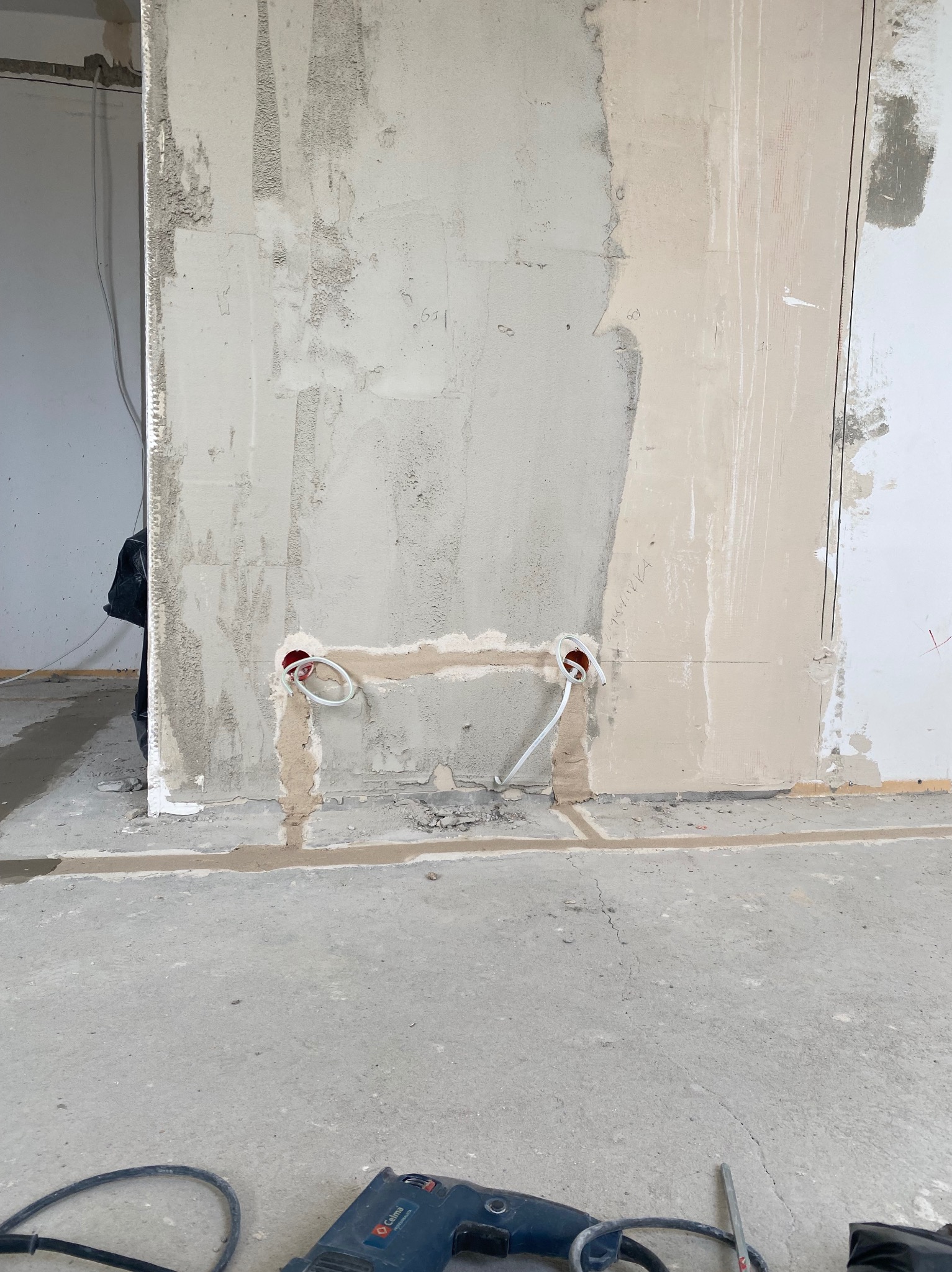 Ściana w trakcie remontu z widocznymi przewodami elektrycznymi w czerwonych puszkach, przygotowana do dalszych prac wykończeniowych. Na podłodze widoczny sprzęt budowlany.