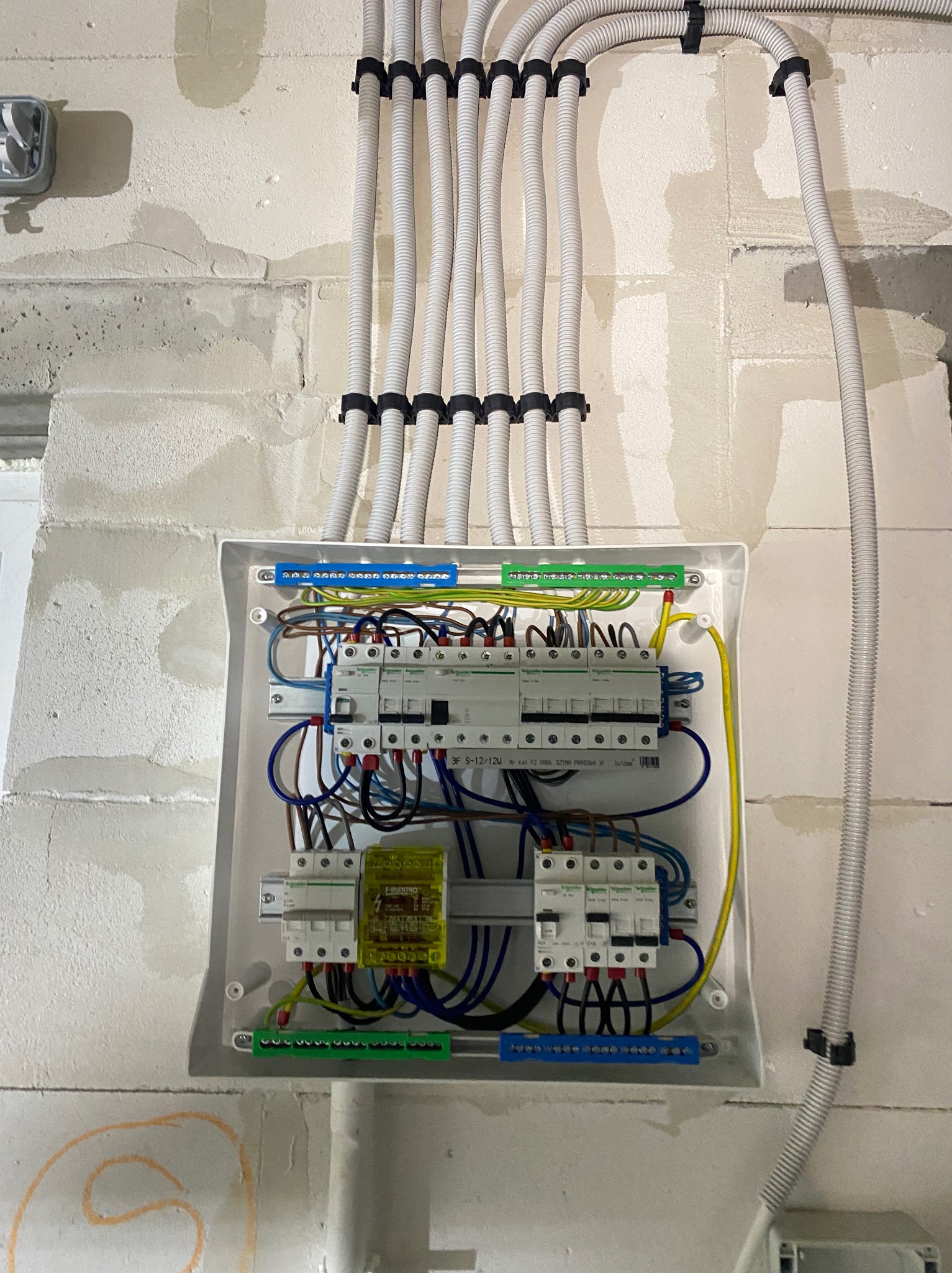 Rozdzielnica elektryczna z przewodami w białej obudowie, zamontowana na ścianie z betonu komórkowego. Widoczne zabezpieczenia i kable w peszlach.