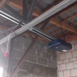 Elektrotech Piotr Wierzchowski - Zamontowany napęd do bramy garażowej z widoczną szyną prowadzącą i elementami mocującymi do sufitu. Surowy mur z bloczków w tle.