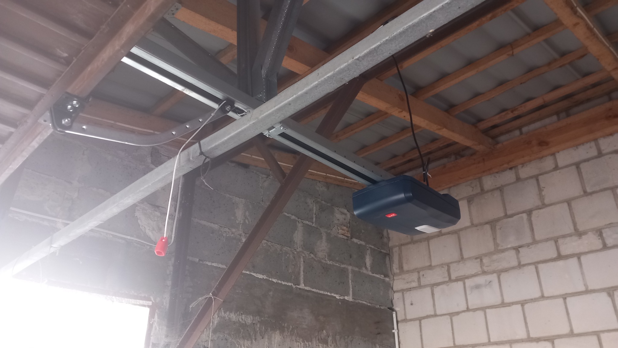Zamontowany napęd do bramy garażowej z widoczną szyną prowadzącą i elementami mocującymi do sufitu. Surowy mur z bloczków w tle.