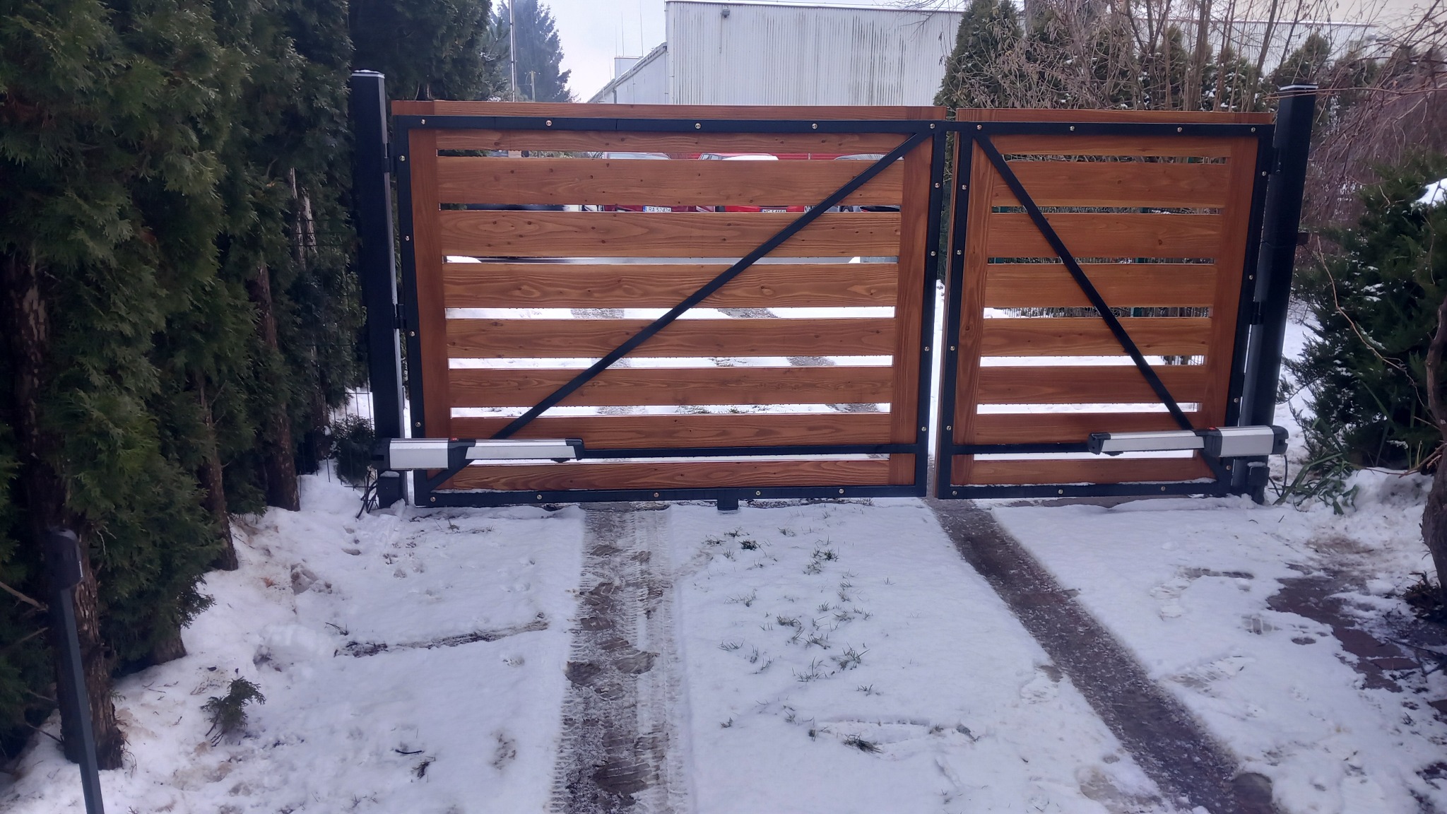 Drewniana brama wjazdowa z automatyką, czarne metalowe wzmocnienia, zimowy krajobraz z zielenią w tle. Widoczne ślady opon na śniegu.