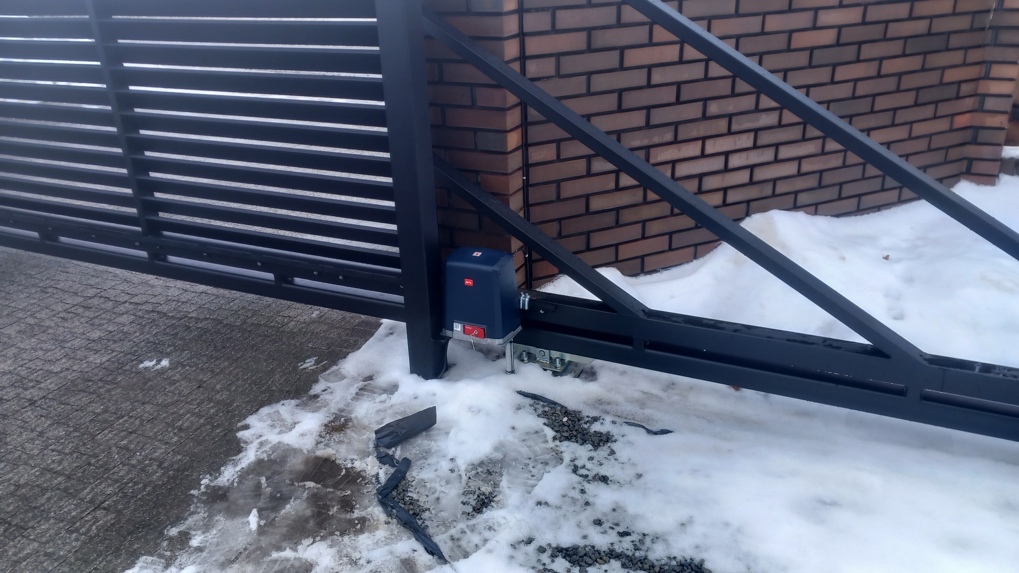 Automatyczna brama przesuwna z napędem, widoczny mechanizm i fragment muru z cegły w tle, częściowo pokryta śniegiem.