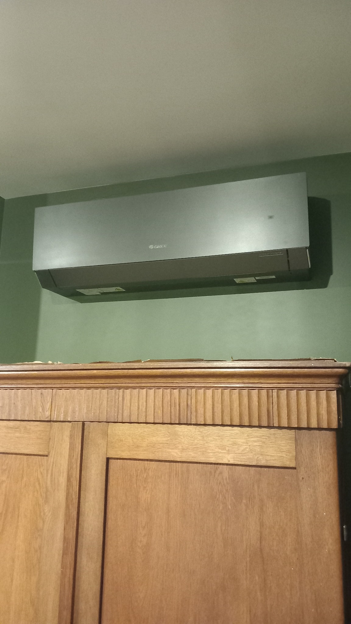 Zamontowany klimatyzator marki Gree na zielonej ścianie, nad drewnianą szafką. Widoczne detale urządzenia i otoczenia. Ujęcie z dołu.
