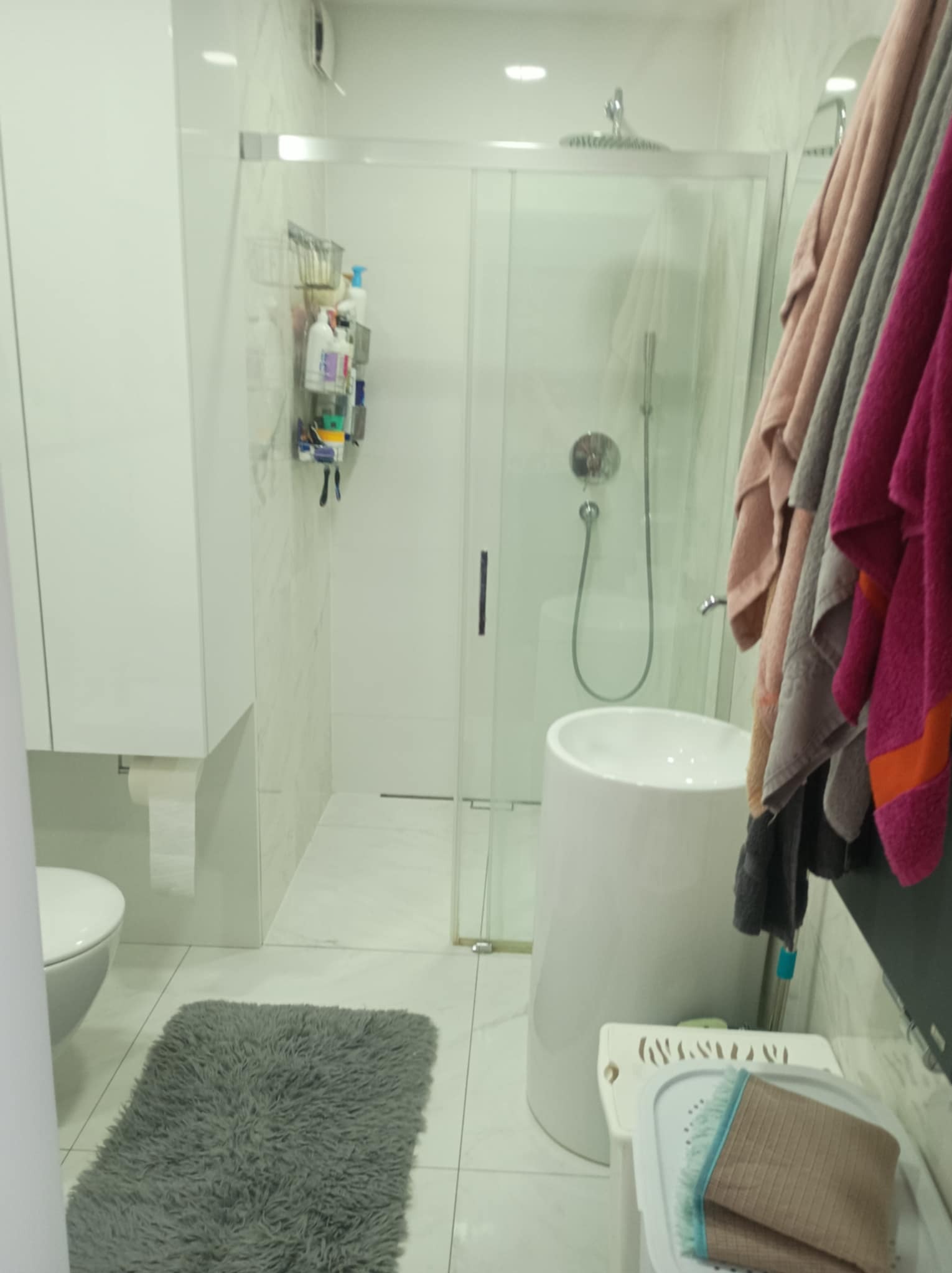 Jasna łazienka z kabiną prysznicową, umywalką na postumencie i szarym dywanikiem. Widoczne ręczniki w odcieniach różu i szarości. Minimalistyczny design, czyste linie.