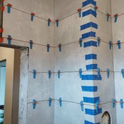 Gol - Ściana w trakcie układania płytek, z użyciem niebieskiej taśmy i pomarańczowych krzyżyków dystansowych. Widoczny otwór na rurę. Szare płytki imitujące beton.