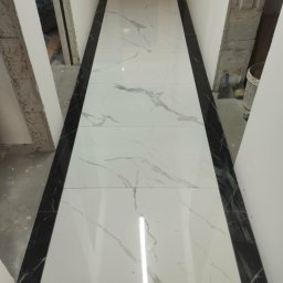 Gol - Białe płytki imitujące marmur z czarnymi obramowaniami, ułożone w korytarzu. Widoczne narzędzia glazurnicze i fragmenty ścian w stanie surowym.