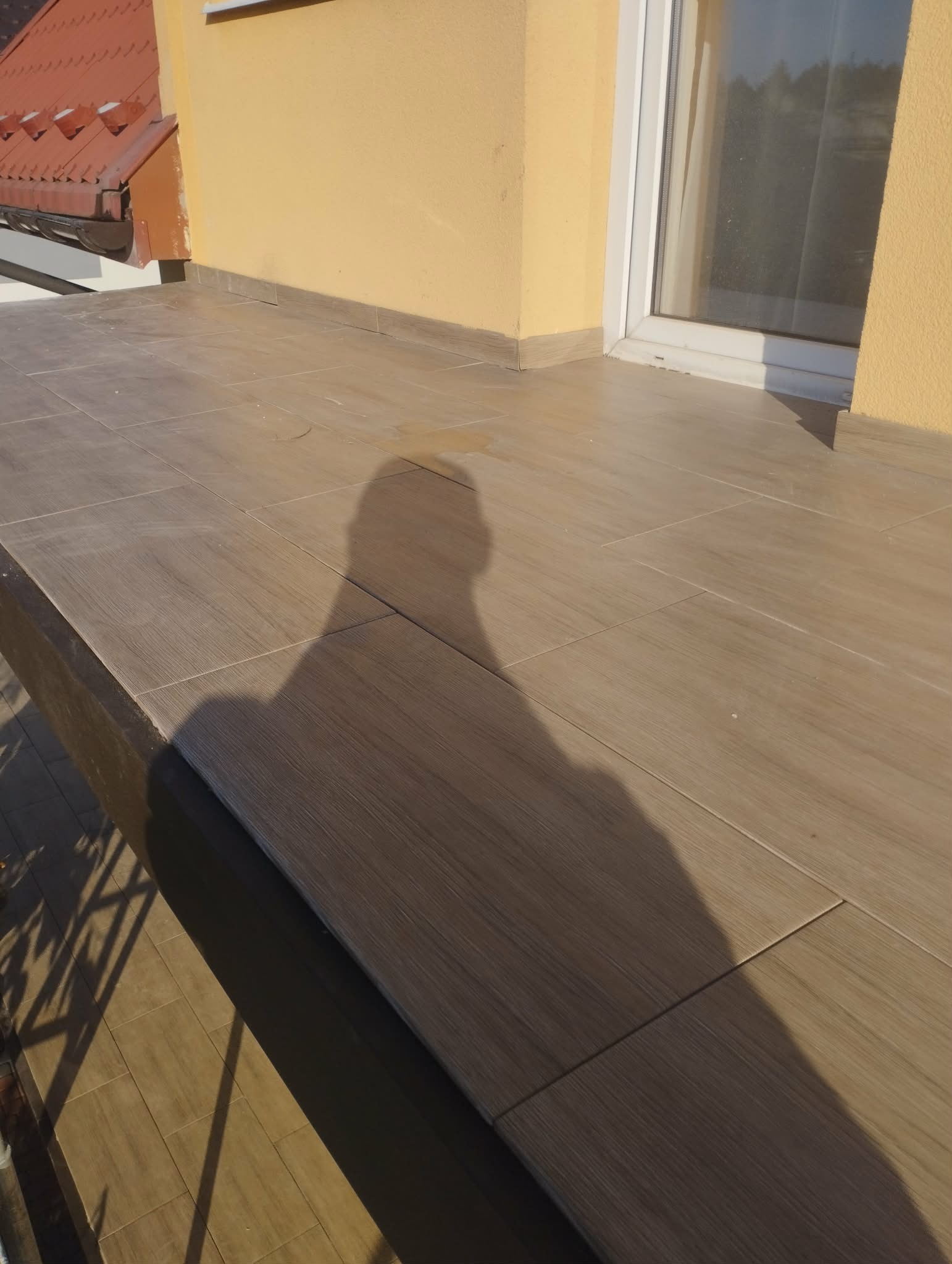 Wykończony balkon z brązowymi płytkami imitującymi drewno, jasna elewacja budynku i cień osoby fotografującej. Widoczne detale wykończenia i fuga.