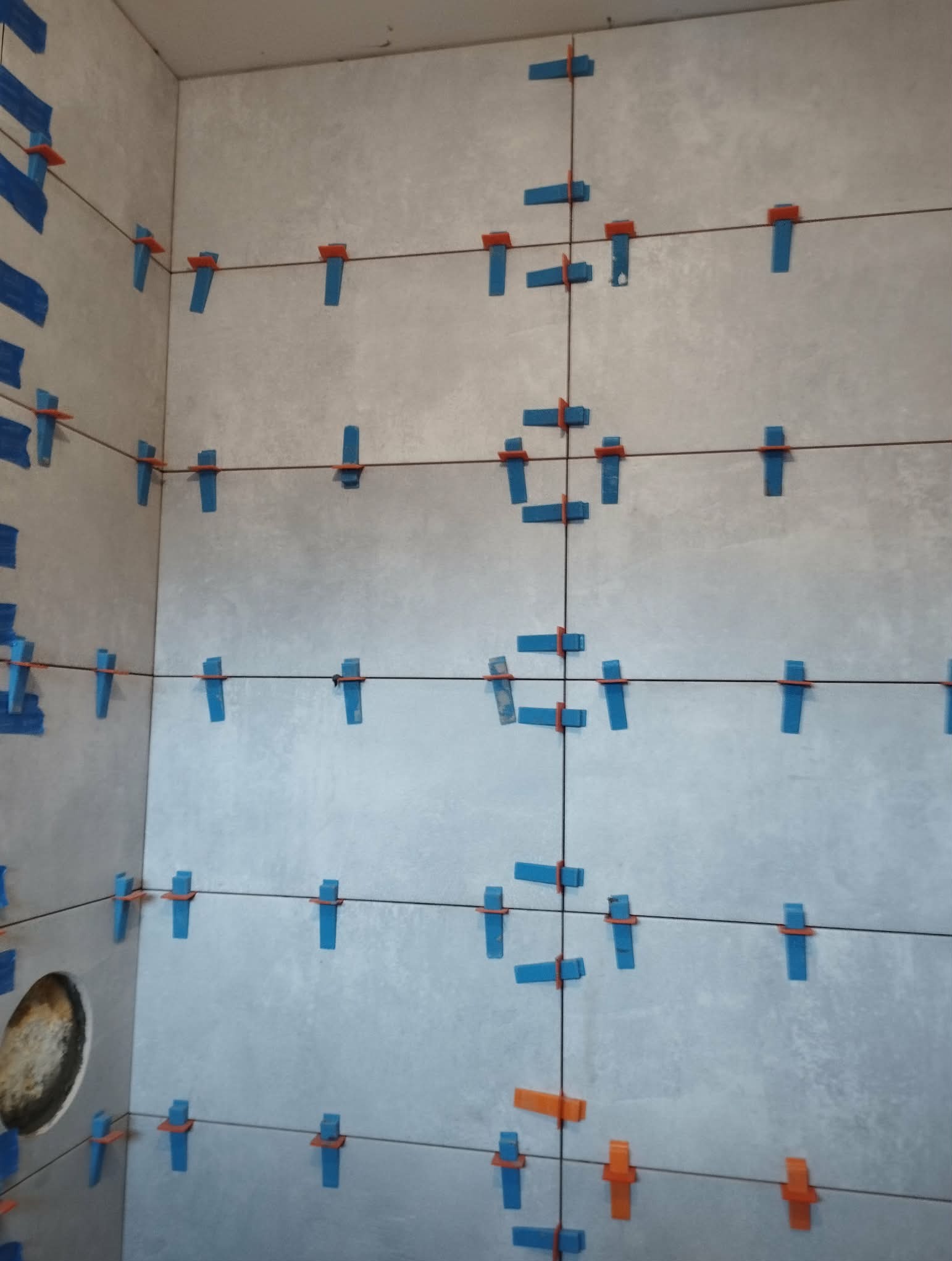 Ściana w trakcie układania płytek, widoczne niebieskie i pomarańczowe klipsy poziomujące, sznurki wyznaczające linie. Fragment otworu w ścianie po lewej.