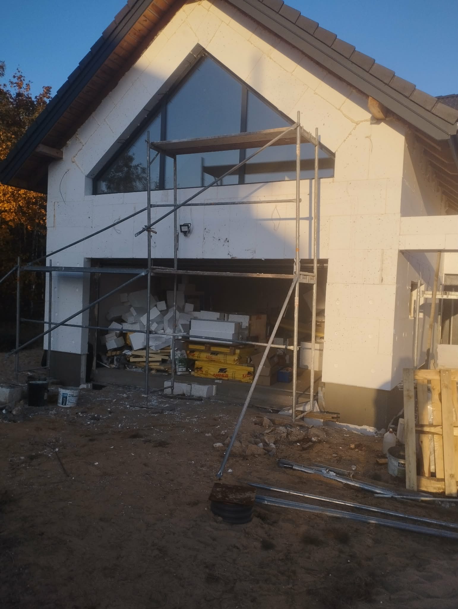 Budowa domu jednorodzinnego z białych bloczków, widoczne rusztowania i materiały budowlane. Okno trójkątne w szczycie. Etap surowy otwarty.