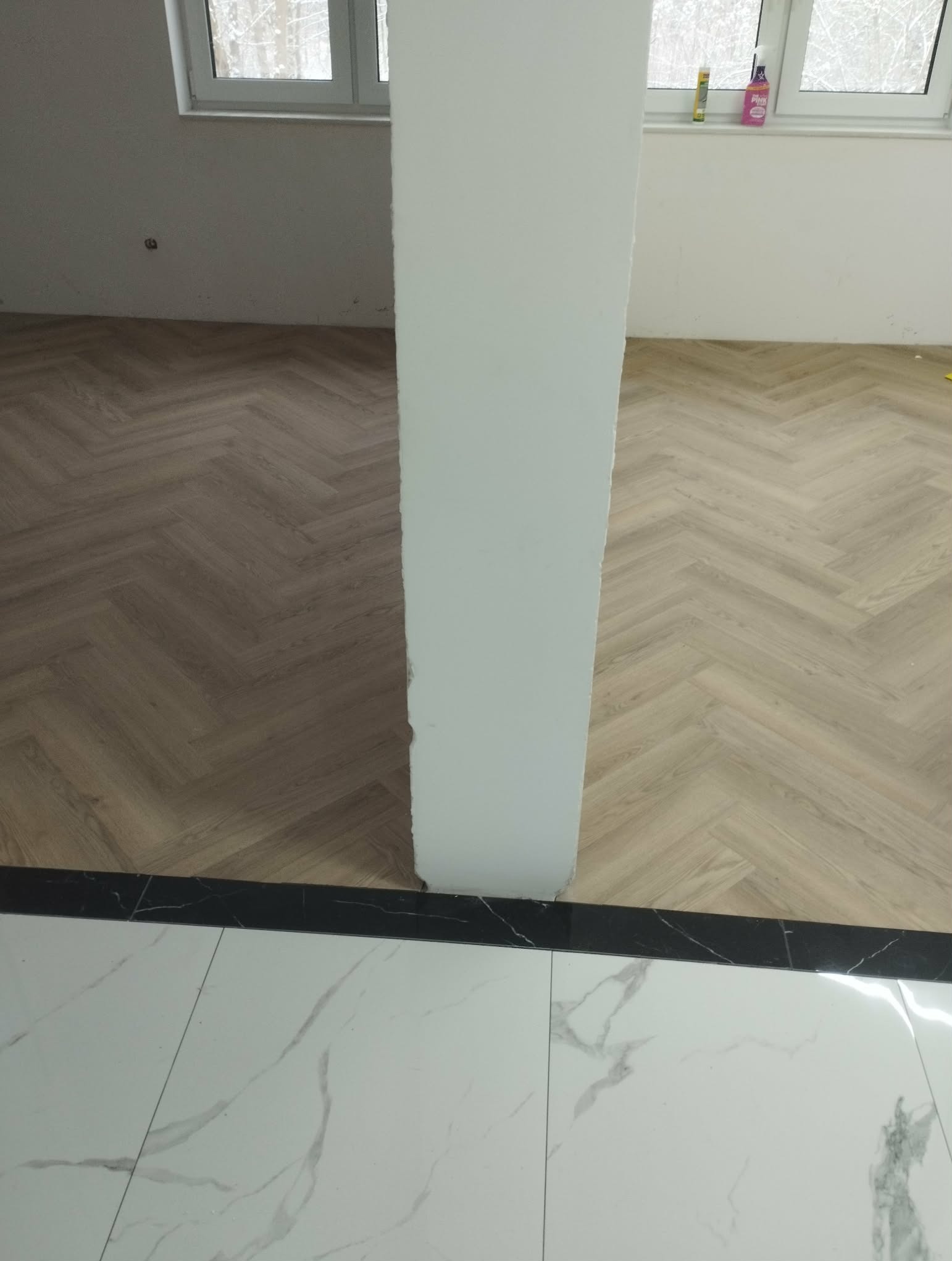 Ukończone podłogi: parkiet w jodełkę w dwóch odcieniach drewna, oddzielone kolumną i czarnym pasem od białych płytek imitujących marmur. Widok na okno.