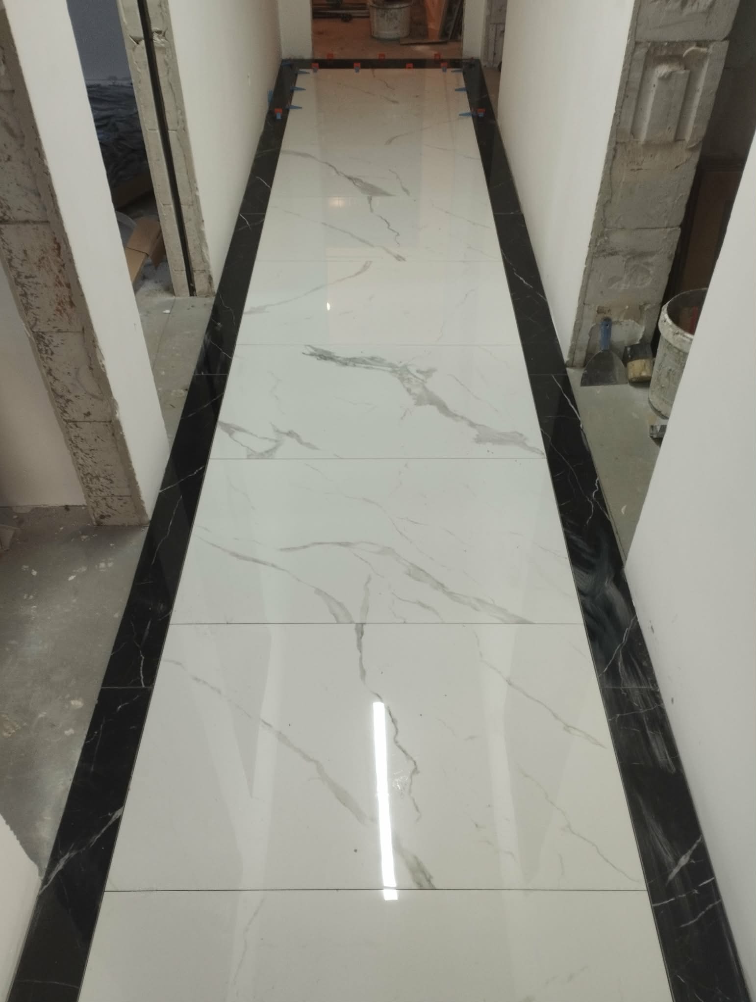 Białe płytki imitujące marmur z czarnymi obramowaniami, ułożone w korytarzu. Widoczne narzędzia glazurnicze i fragmenty ścian w stanie surowym.