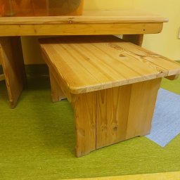 WOODEN ART SPÓŁKA Z OGRANICZONĄ ODPOWIEDZIALNOŚCIĄ - Drewniany stół z ławką w jasnym odcieniu, widoczne słoje drewna. Na stole przezroczysty pojemnik z akcesoriami. Podłoga w odcieniach zieleni i żółci.