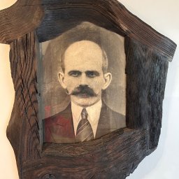 WOODEN ART SPÓŁKA Z OGRANICZONĄ ODPOWIEDZIALNOŚCIĄ - Portret mężczyzny z wąsami w unikatowej, ręcznie robionej ramie z ciemnego drewna o wyraźnej strukturze słojów, wiszący na białej ścianie.
