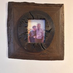 WOODEN ART SPÓŁKA Z OGRANICZONĄ ODPOWIEDZIALNOŚCIĄ - Drewniana rama na zdjęcie z wyraźną fakturą drewna i okrągłym otworem, wisząca na jasnej ścianie. W ramie widoczne zdjęcie portretowe.