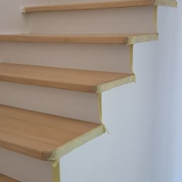 WOODEN ART SPÓŁKA Z OGRANICZONĄ ODPOWIEDZIALNOŚCIĄ - Jasne, drewniane stopnie schodów zabezpieczone taśmą malarską na białej ścianie. Widok z dołu, ukazujący detale wykończenia i przygotowania do malowania.