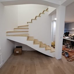 Spiralne schody z jasnego drewna w trakcie montażu, widoczne zabezpieczenia na stopniach. W tle pomieszczenie z narzędziami i elementami wykończenia wnętrz.