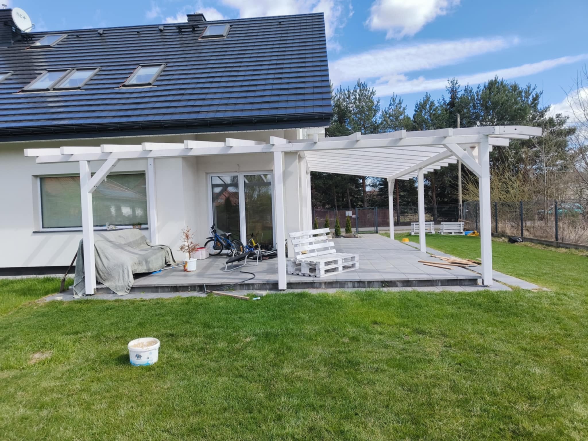 Biała pergola z drewnianą konstrukcją na tarasie z szarych płyt, z widokiem na trawnik i dom z ciemnym dachem w tle, w słoneczny dzień. Widoczny fragment mebli ogrodowych.