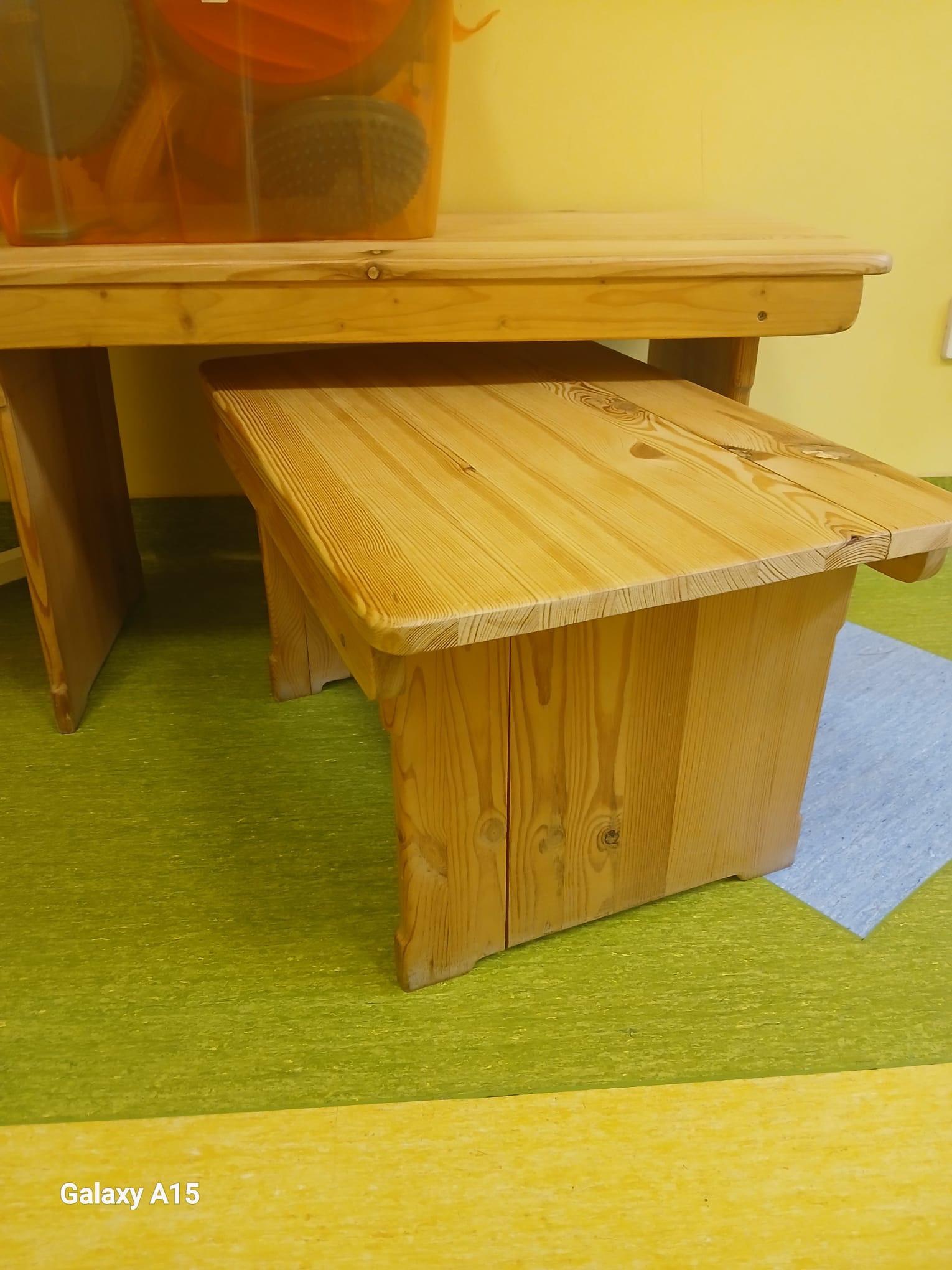 Drewniany stół z ławką w jasnym odcieniu, widoczne słoje drewna. Na stole przezroczysty pojemnik z akcesoriami. Podłoga w odcieniach zieleni i żółci.