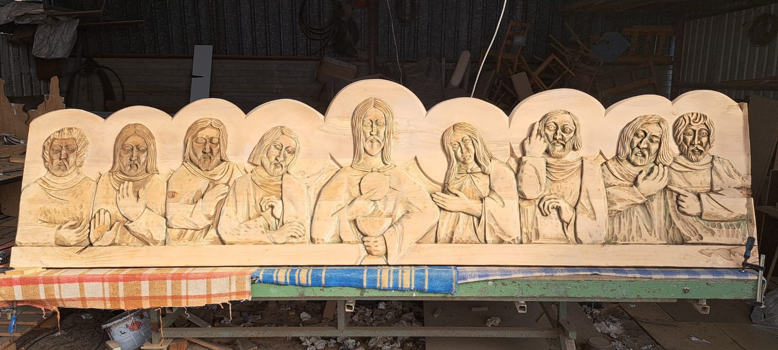 Ręcznie rzeźbiona płaskorzeźba z jasnego drewna przedstawiająca scenę Ostatniej Wieczerzy, umieszczona na stole warsztatowym w pracowni stolarskiej.