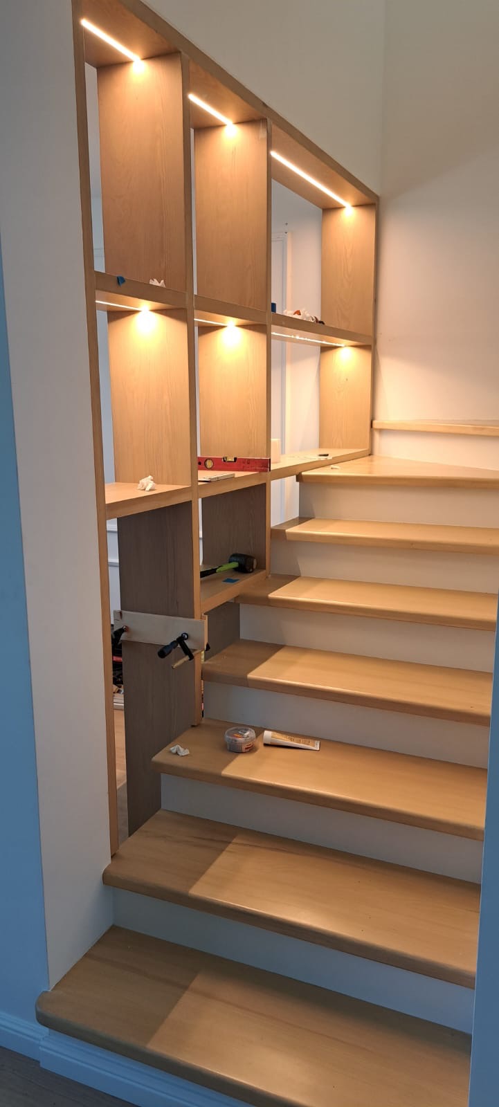 Drewniane schody z regałem, oświetlenie LED. Widoczne narzędzia: poziomica, młotek. Jasne drewno, minimalistyczny design. Ukończony projekt z elementami wykończeniowymi.