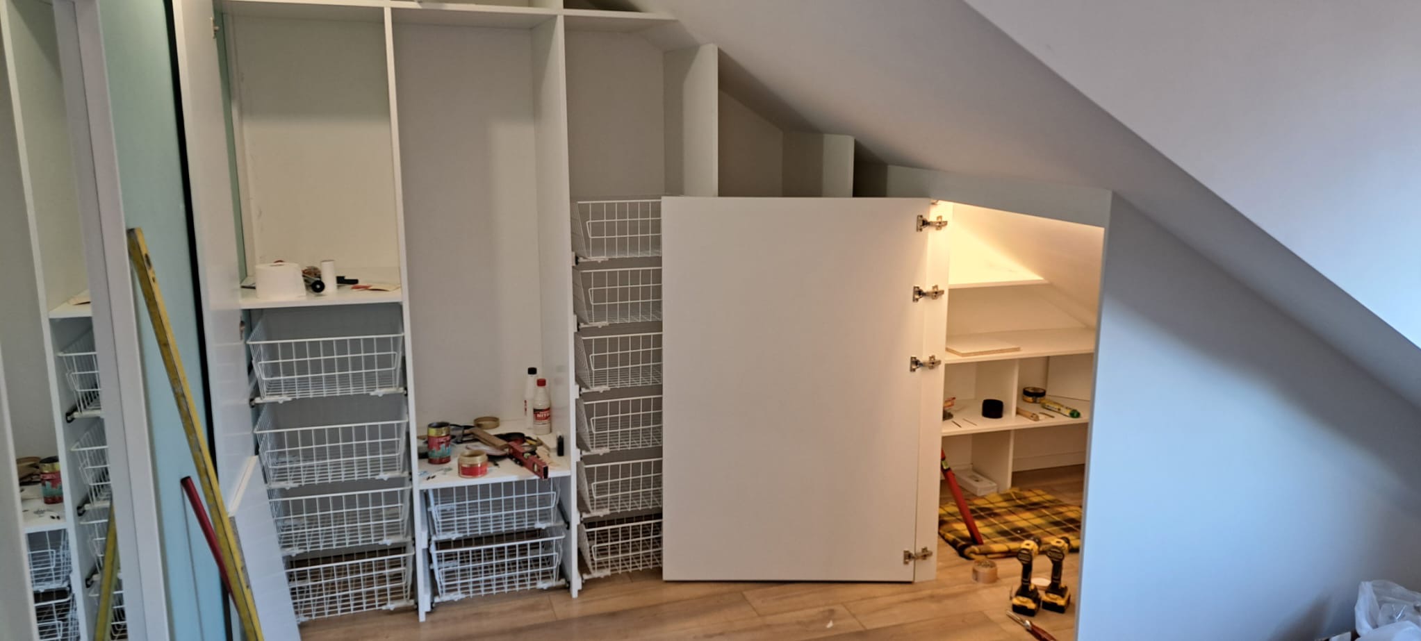 Biała zabudowa garderoby na poddaszu ze stalowymi koszami i otwartą szafką z półkami. Widoczne narzędzia i poziomica.