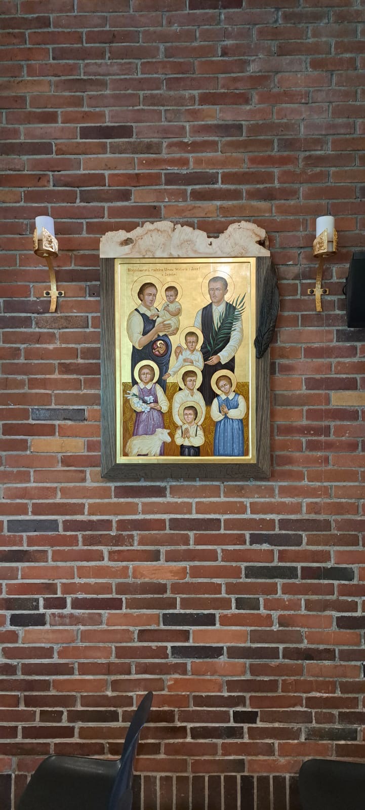 Ikona Świętej Rodziny w drewnianej ramie, zawieszona na ceglanej ścianie, oświetlona przez kinkiety ze świecami. Ujęcie z dołu, widoczny fragment krzesła.