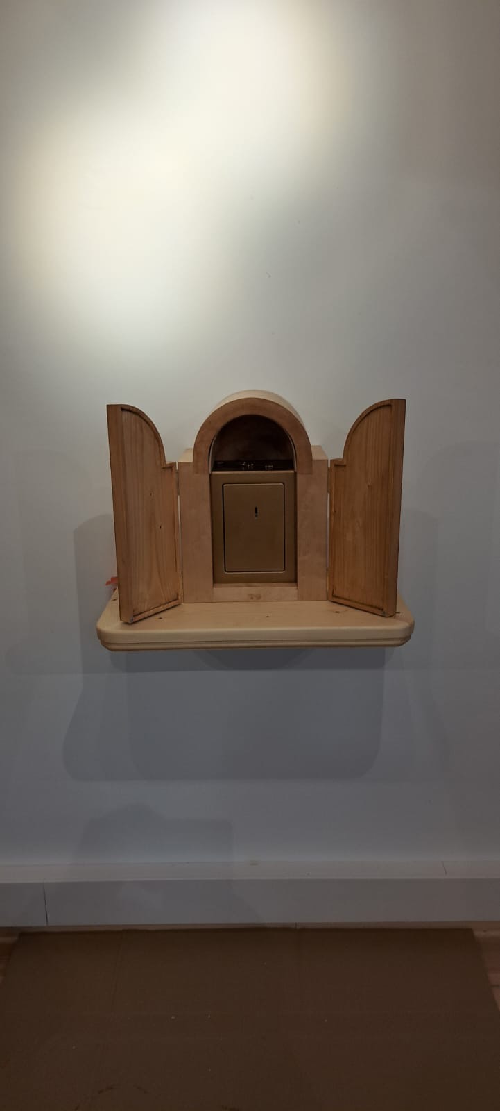 Drewniana szafka ścienna z otwartymi drzwiczkami, zamontowana na półce. Naturalne drewno, minimalistyczny design, oświetlenie górne. Widok z przodu, na jasnym tle ściany.