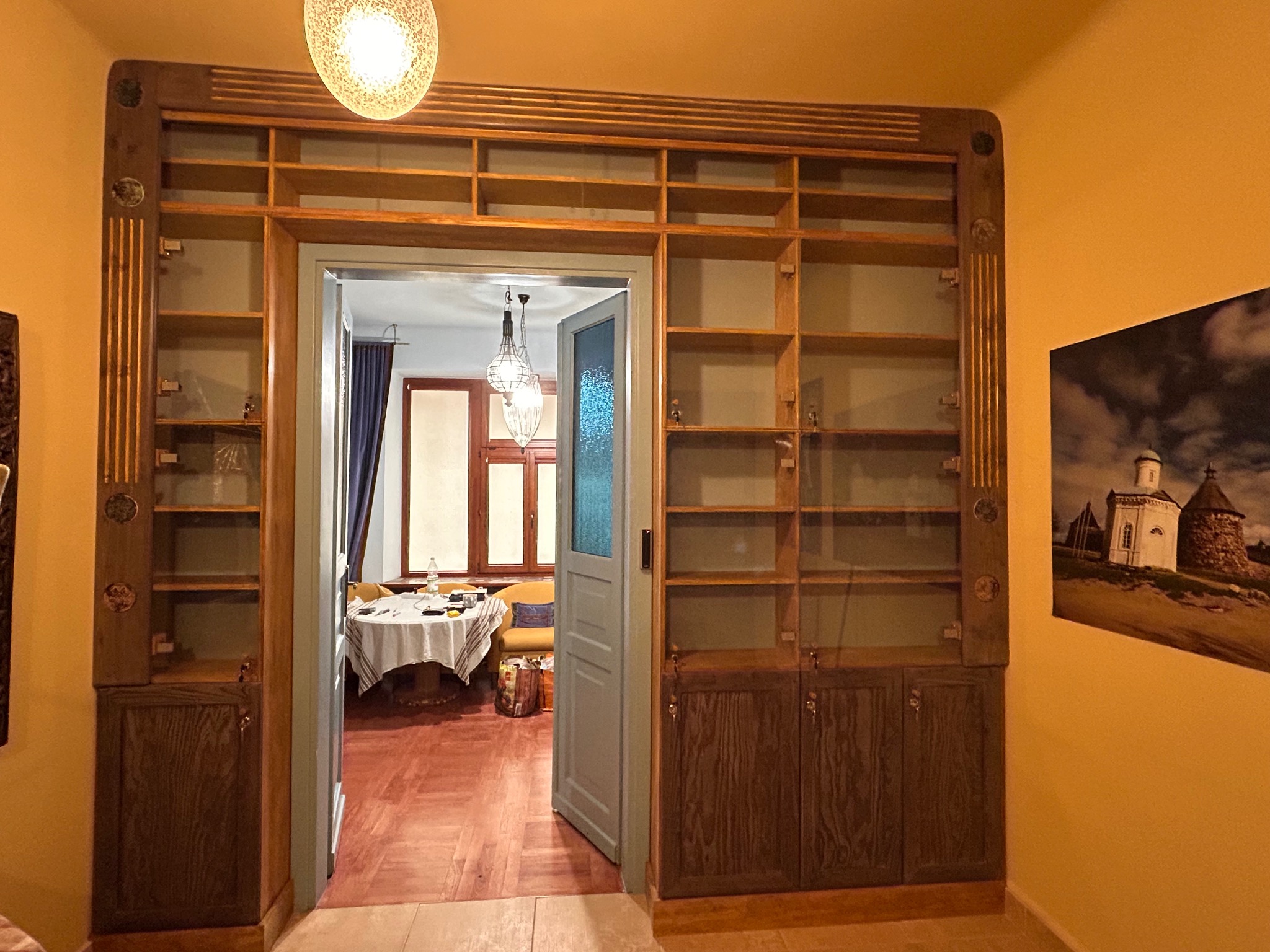 Drewniana zabudowa regałowa z szafkami i otwartymi półkami, wbudowana we framugę drzwi. W tle widoczny pokój z nakrytym stołem i oknem.