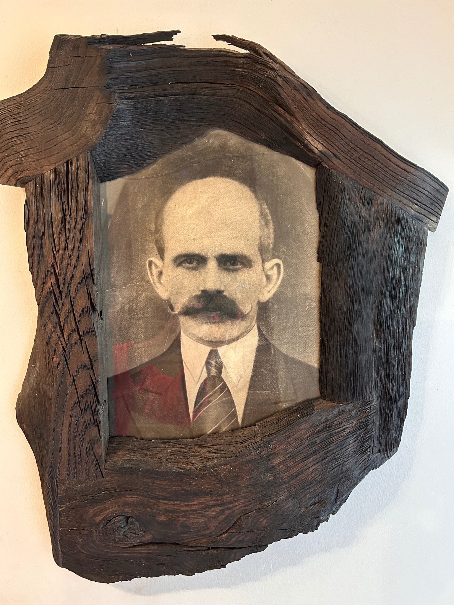 Portret mężczyzny z wąsami w unikatowej, ręcznie robionej ramie z ciemnego drewna o wyraźnej strukturze słojów, wiszący na białej ścianie.