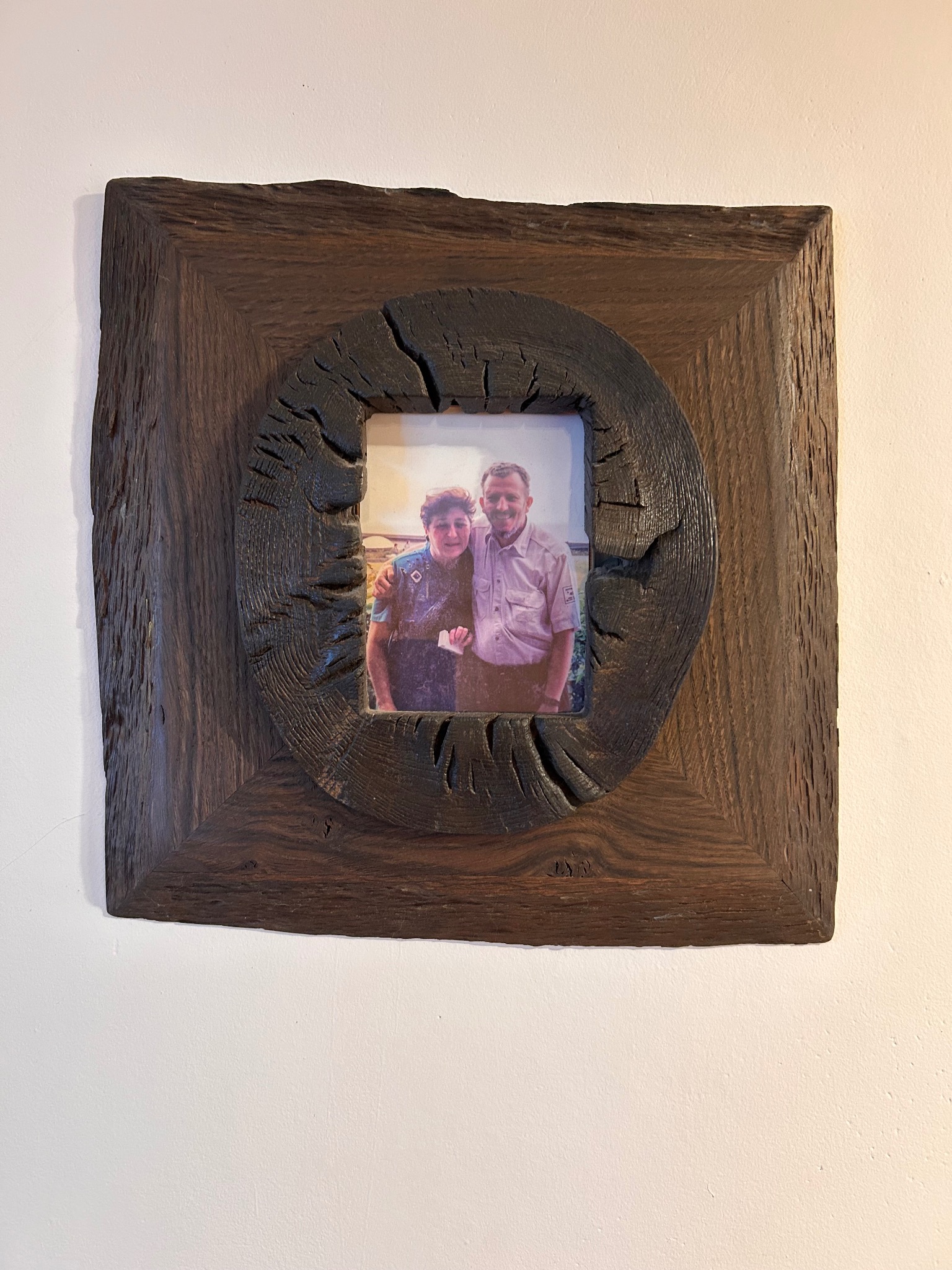 Drewniana rama na zdjęcie z wyraźną fakturą drewna i okrągłym otworem, wisząca na jasnej ścianie. W ramie widoczne zdjęcie portretowe.