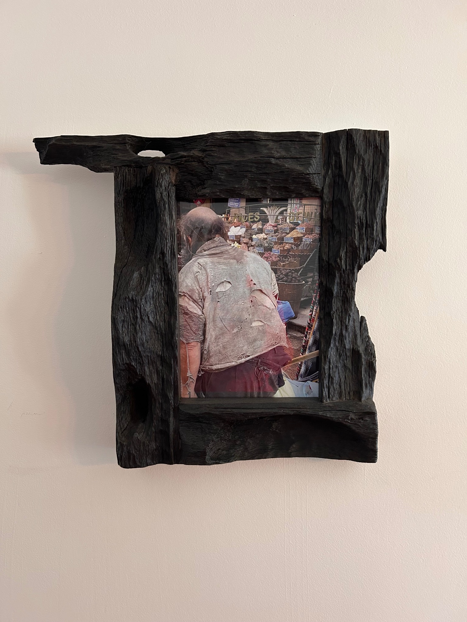 Nietypowa rama z ciemnego drewna o nieregularnym kształcie, wisząca na jasnej ścianie, otacza fotografię przedstawiającą scenę z targu.