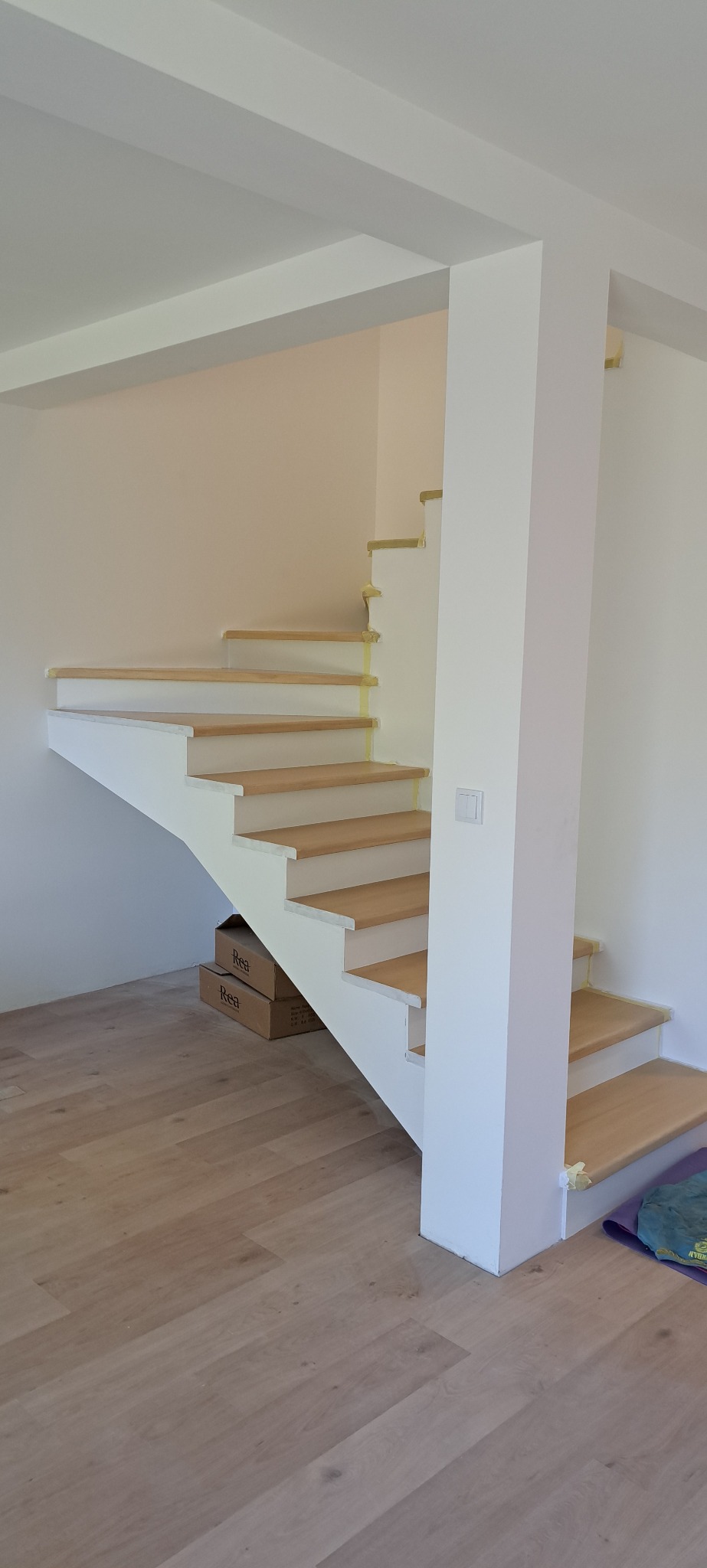 Nowoczesne schody drewniane z białymi podstopnicami i drewnianymi stopniami, zabezpieczone taśmą malarską, w jasnym wnętrzu. Widoczne kartony i podłoga z paneli.
