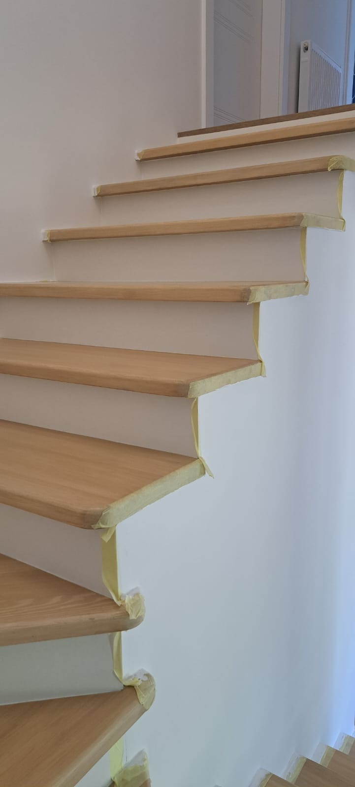 Jasne, drewniane stopnie schodów zabezpieczone taśmą malarską na białej ścianie. Widok z dołu, ukazujący detale wykończenia i przygotowania do malowania.