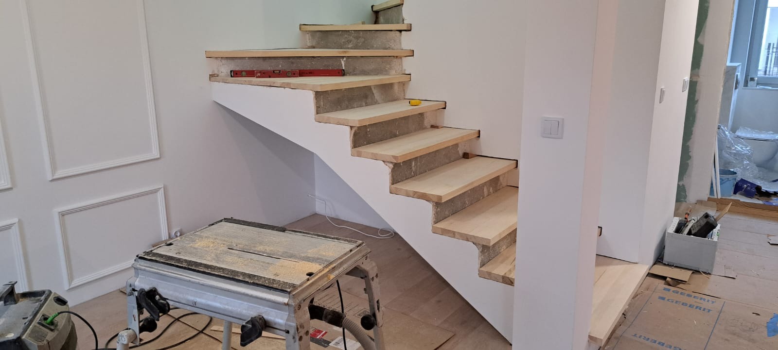 Wnętrze w trakcie montażu drewnianych schodów z poziomnicą i piłą stołową. Widoczne surowe stopnie i białe ściany z dekoracyjnymi panelami.