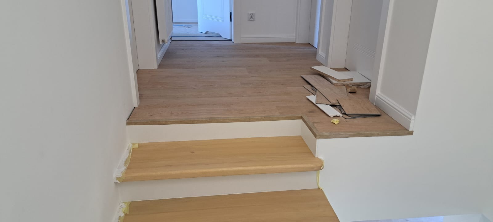 Montaż drewnianych schodów w jasnym wnętrzu. Widoczne fragmenty schodów z jasnego drewna, podłoga z paneli i białe ściany. Etap prac wykończeniowych.