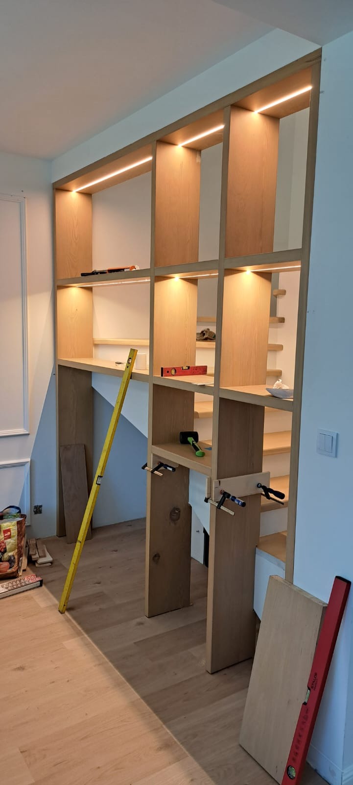 Drewniana zabudowa regałowa z oświetleniem LED w trakcie montażu, widoczne narzędzia i poziomice. Konstrukcja wkomponowana w przestrzeń schodów.
