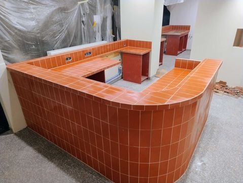 Wykończona, pomarańczowa lada barowa z płytek w kształcie litery U, z widocznymi fragmentami instalacji i resztkami materiałów budowlanych w tle.