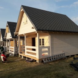 Monstar - Trzy identyczne, jasne domki szkieletowe z ciemnymi dachami, ustawione w rzędzie. Na pierwszym planie pracownik w trakcie wykonywania prac wykończeniowych.