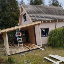 Monstar - Wznoszenie domu szkieletowego: drewniana konstrukcja z częściowo ułożonym dachem, drabina oparta o budynek, widoczne elementy wykończeniowe i narzędzia na trawie.