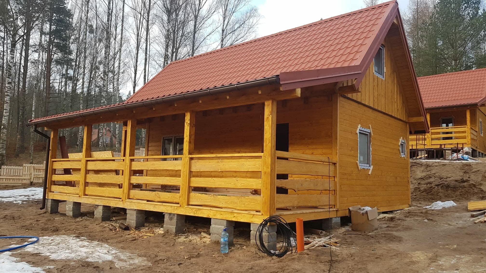 Surowy, drewniany dom szkieletowy z czerwonym dachem na betonowych fundamentach, z widoczną werandą i elementami instalacji na ziemi. W tle drzewa.