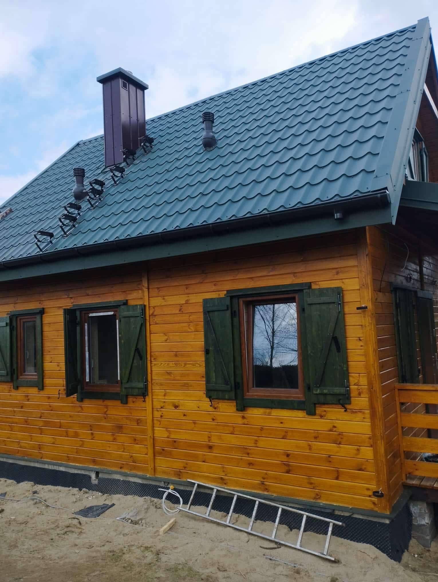 Żółty dom szkieletowy z zielonym dachem i okiennicami, widoczny fragment fundamentu i drabina oparta o ścianę. Charakterystyczny komin i wentylacja na dachu.