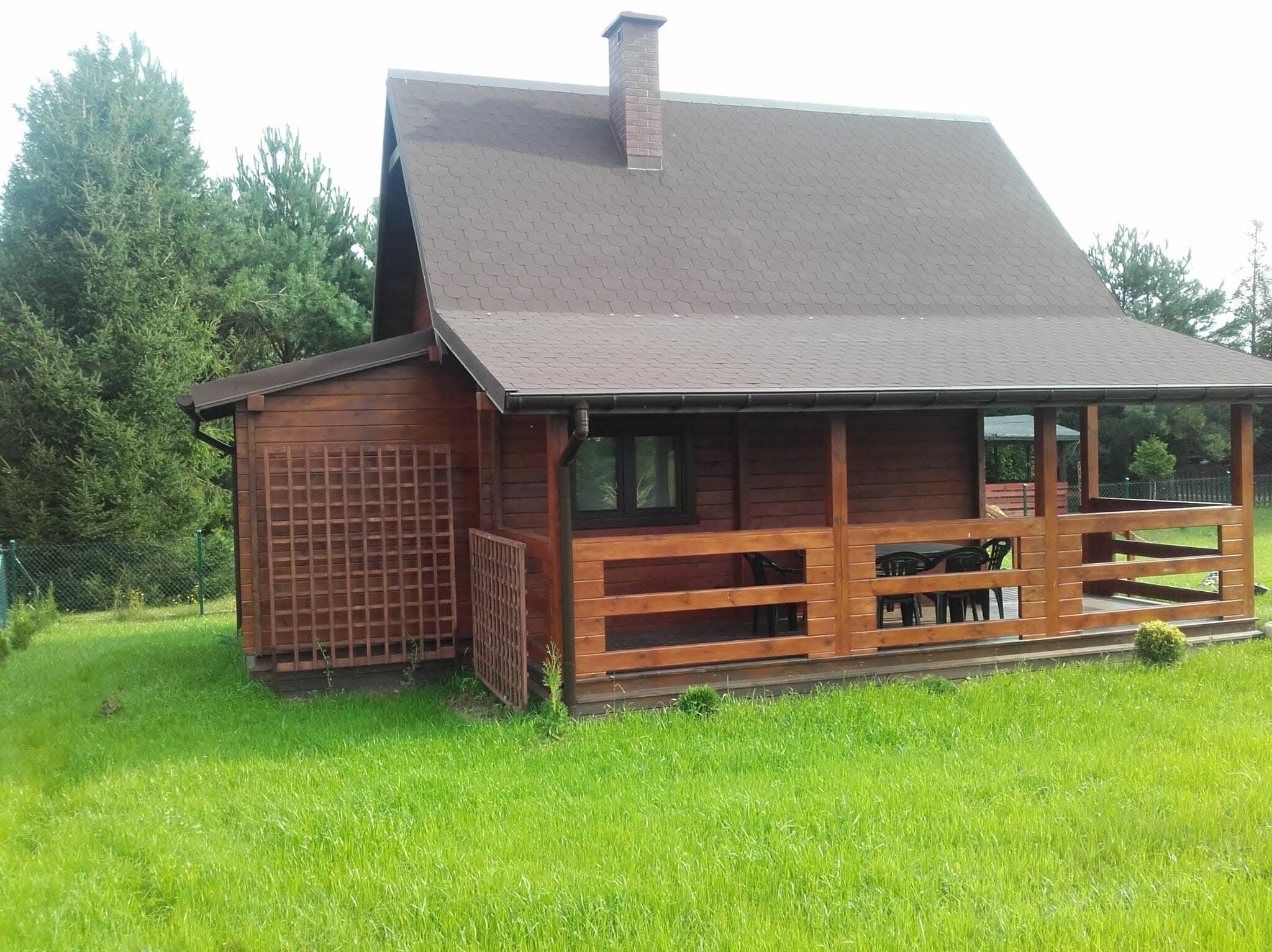Drewniany dom z brązowym dachem i kominem, otoczony zielonym trawnikiem i drzewami. Widoczna weranda z meblami ogrodowymi.