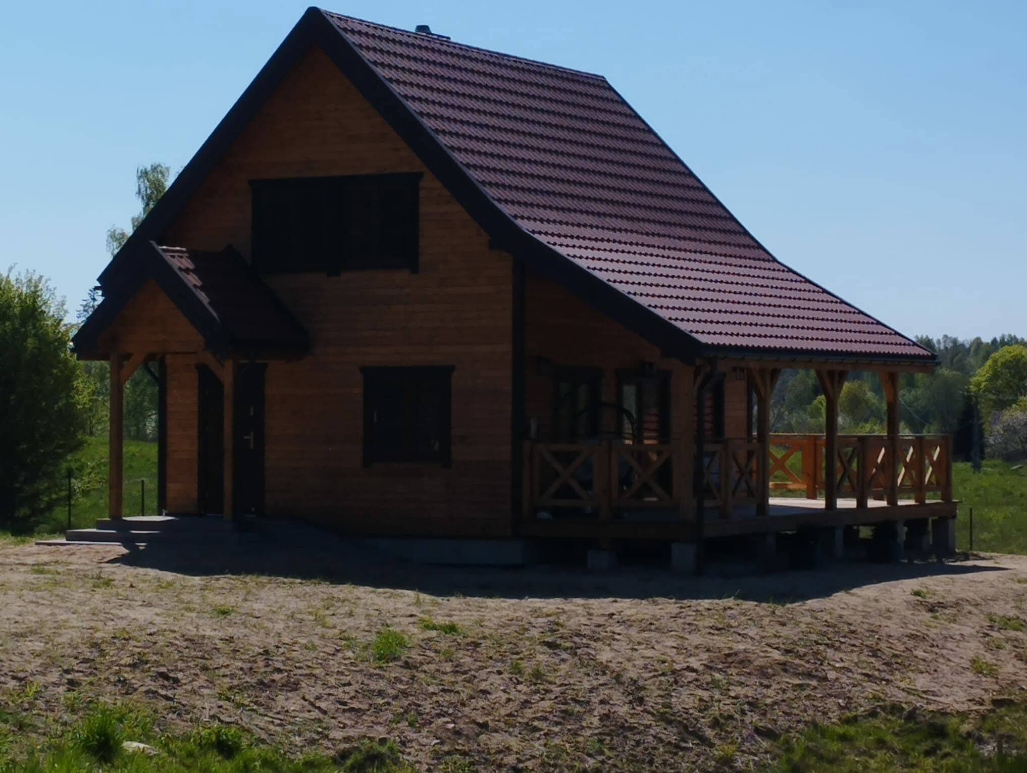 Gotowy, brązowy dom szkieletowy z ciemnoczerwoną dachówką, widoczny z przodu, z drewnianym tarasem i schodami wejściowymi, na tle zieleni i błękitnego nieba.